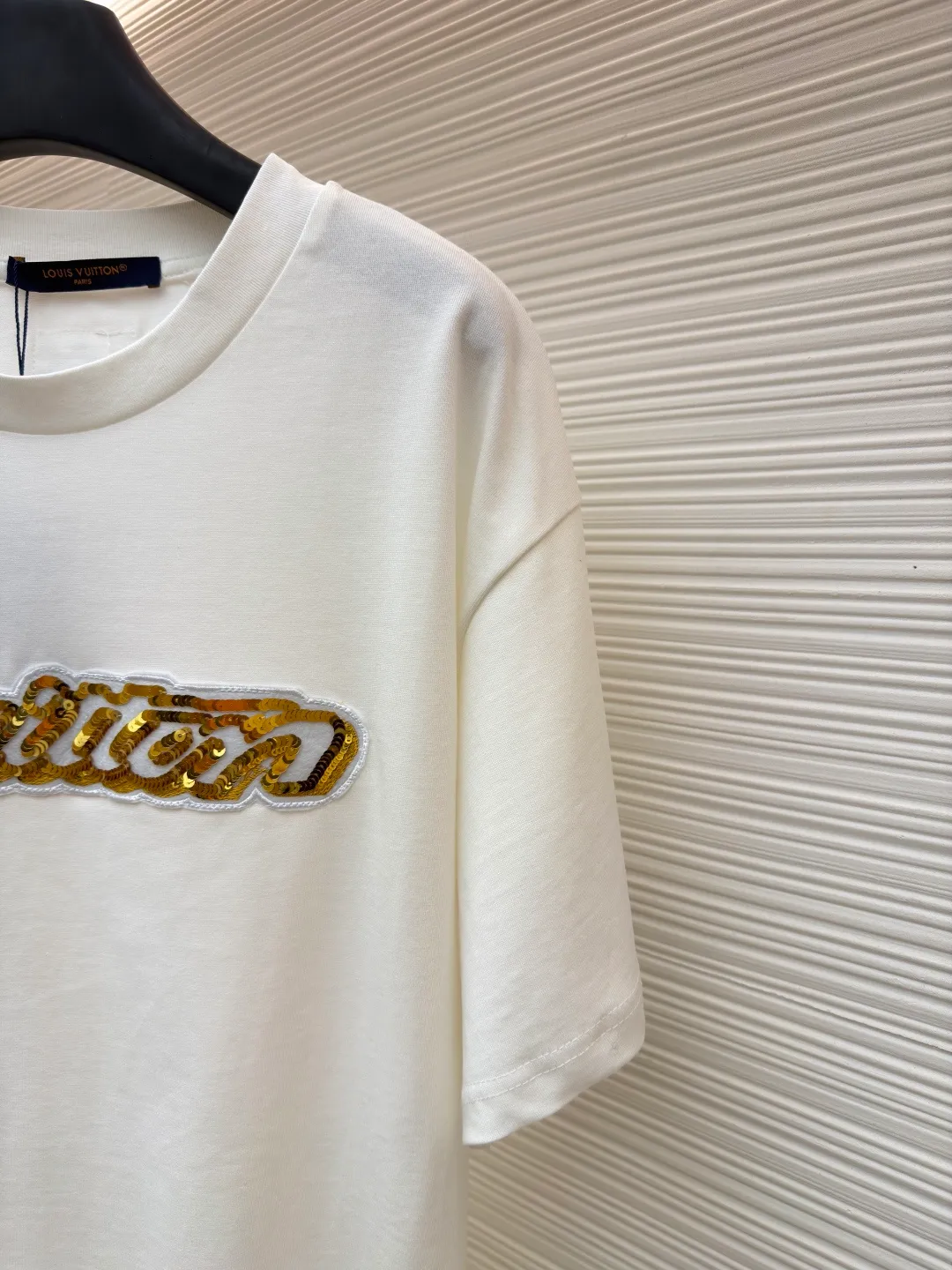 LOUIS VUITTON cotton round neck embroidered Vuitton signature logo short sleeves ●Original version: purchased and developed for 12300 ●Item number: 1682272-A10 ●Fabric: cotton fabric ●Technology: embroidery production ●Color: white ●Version: fitted version ●Weight: about 270 grams ●Size: S-M-L-XL ●Description: This white short-sleeved T-shirt is made of high-count combed cotton fabric and has been treated with pre-washing technology. It has a soft and skin-friendly touch and is not easy to deform. The silhouette of the shirt is a loose shoulder design, which meets the needs of modern casual wear. The core decoration area on the chest uses hand-beading and bronzing technology, and uses metallic gold sequins and embroidery threads intertwined to outline a three-dimensional letter pattern. The sequins are layered and inlaid, and each one is manually positioned and fixed by craftsmen to ensure a regular texture and uniform gloss. The edges of the letters are sewn with matte silk thread to form a visual contrast between gold and matte, while reinforcing the decorative structure to avoid falling off during wear. The neckline uses double-needle hemming technology and is matched with ribbed cuffs of the same fabric. It is durable and fits the neck. The overall craftsmanship combines handicraftsmanship with modern clothing technology, and the details show exquisite texture. The overall wiring is straight and delicate, the fabric is comfortable and soft, and the color is consistent without color difference. It can be matched with the same style to create a fashionable look and control various occasions. Customized accessories, complete with three standards, same style for men and women!